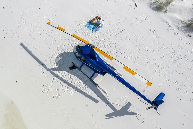 A small helicopter parked on white sand, seen from above, a picnic blanket laid out beside it
