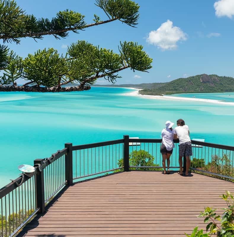 Two walkers at the rail of the Tongue Point lookout, hoop pines overhead and the swirl of Hill Inlet below